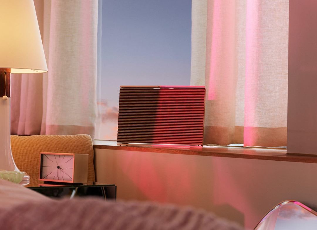 Brown and black speaker sitting on a windowsill with a lamp and clock visible in a warmly lit room against a sky background.