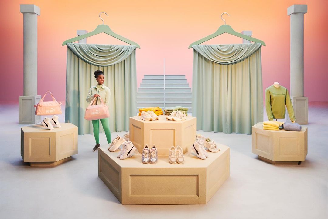 A young Black woman in sportswear holds a pink Puma duffel bag in a colorful studio with display tables of Puma shoes and apparel.