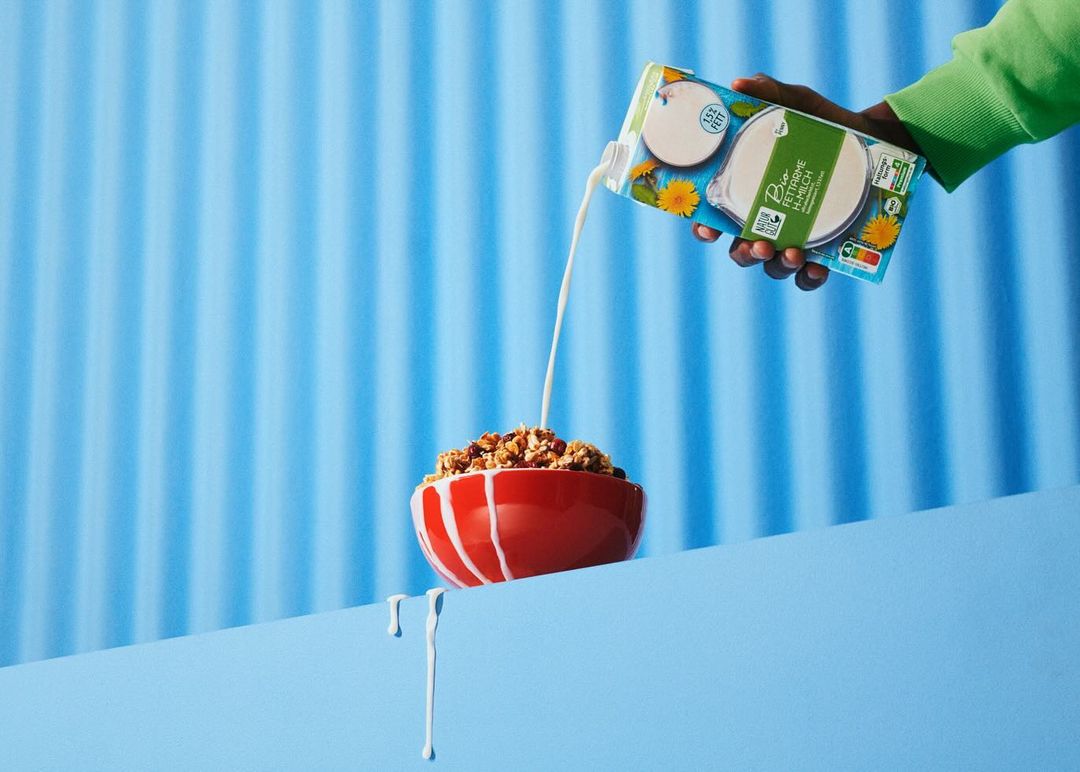 Cereal is being poured from a carton into a bowl by a person wearing a green sweater, set against a vibrant blue background.