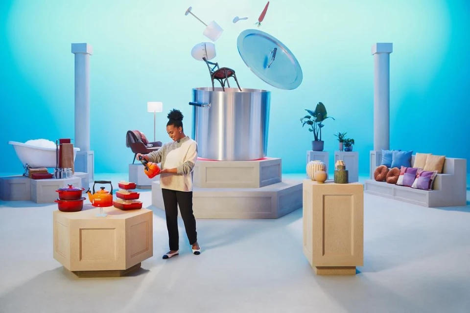 Woman pouring a kettle in front of a giant metal pot floating household objects on a blue set with pedestals and pillars.