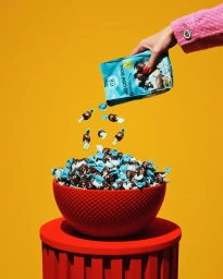 Hand pouring small wrapped candies from a blue bag into a red patterned bowl resting on a red pedestal against a yellow background.