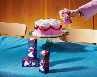 Hand in pink jacket grabbing a slice of frosted pink cake beside blue cartons on a blue tablecloth