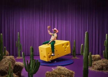A Black man in a pink cowboy hat sits astride a giant yellow cheese block on a stage with a purple curtain backdrop and scattered cacti.
