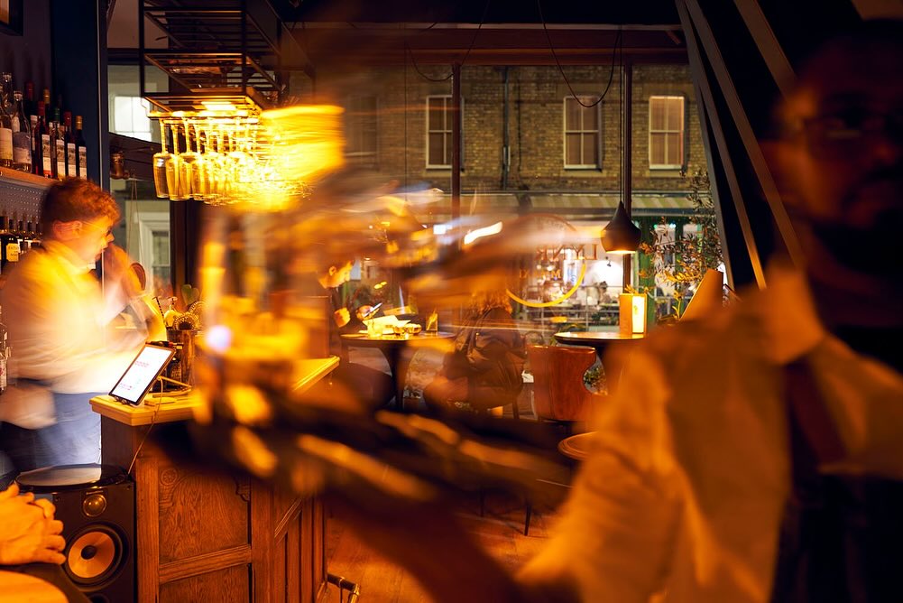 Blurred view inside a bar with bartenders, customers at tables, warm lighting, and a street visible through a window.