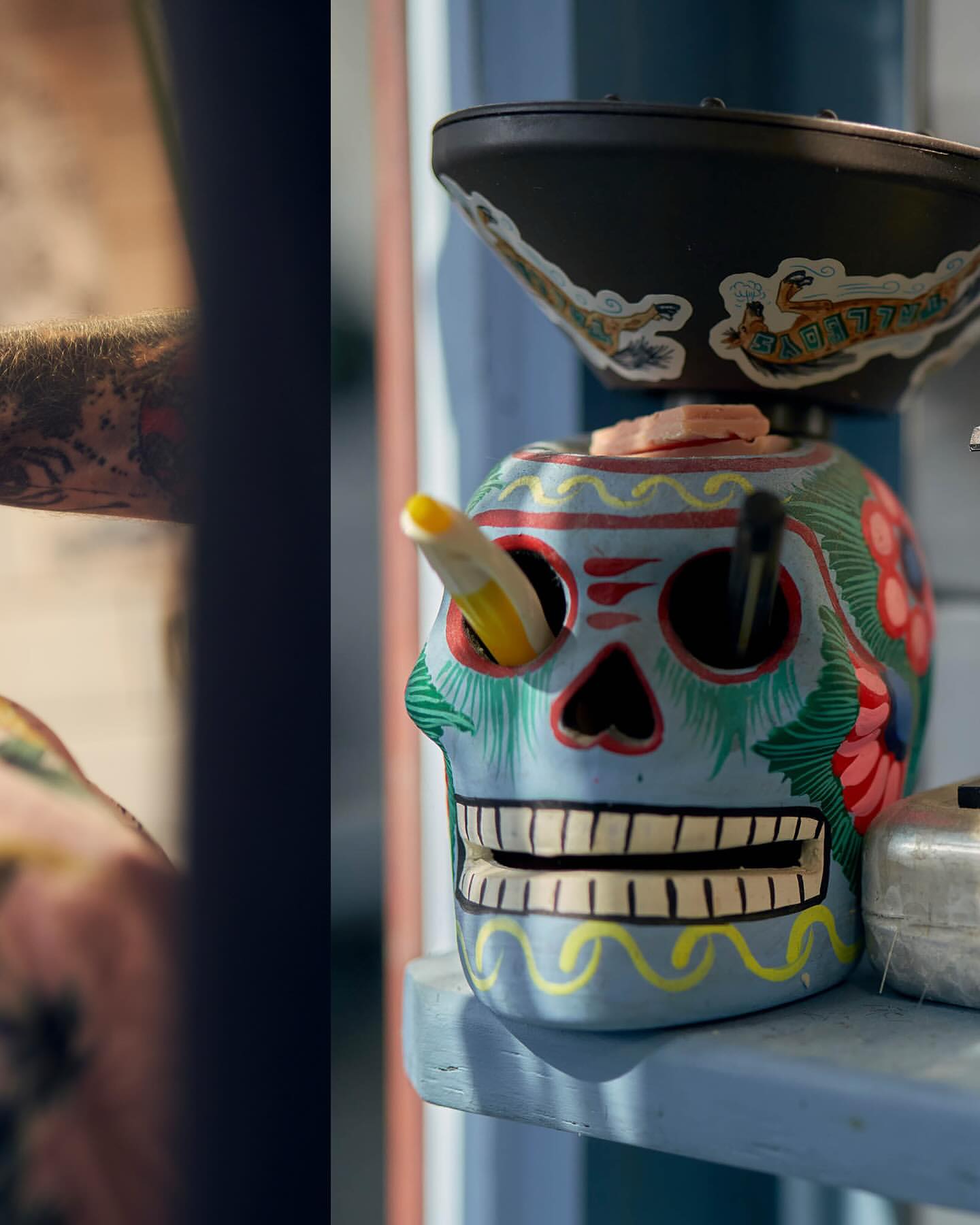 Close-up of a colorful skull-shaped object with a black bowl on top, pens inside. Sitting on a light blue shelf, whimsical.