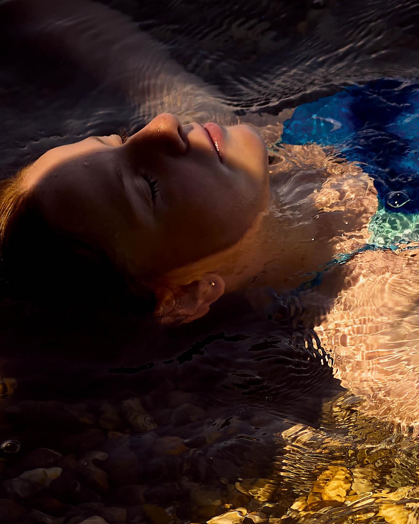 Woman floats on her back in shallow water, face tilted up. Sunlight catches ripples in the water and reflects off the rocks below.