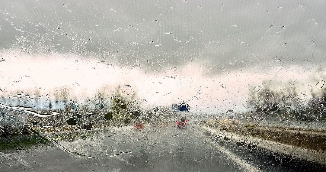 Raindrops on a car windshield obscure the view of the wet road, trees, and overcast sky, creating a moody, blurred, and atmospheric scene.