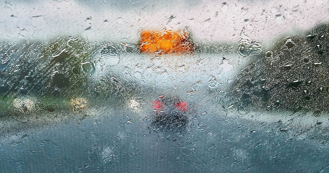 Raindrops on a car windshield obscure the view of a vehicle on a highway, with an orange sign visible in the blurred background.