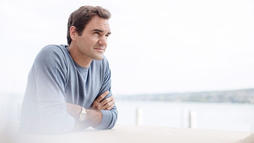 Portrait of an adult male with his arms crossed wearing a sweater. The background includes a body of water and a pale sky.