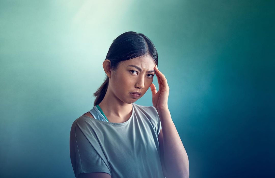 Close-up of an Asian woman with a hand on her forehead, a furrowed brow, and a serious expression, set against a blue-green gradient backdrop.
