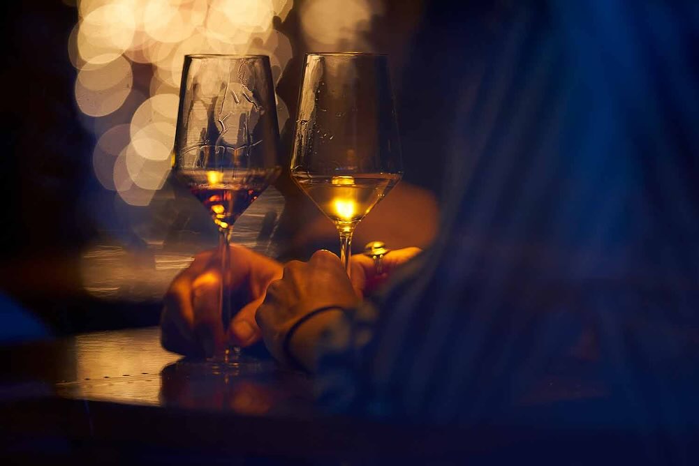Close-up shot of two wine glasses with liquid on a wood surface, against a blurry background with warm lights, creating a moody atmosphere.