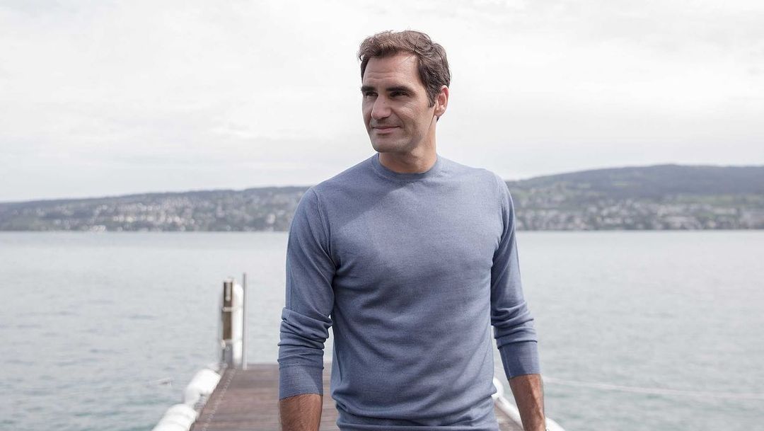 Portrait of adult male in a blue long-sleeve shirt standing on a wooden dock with lake and distant hills in the background.