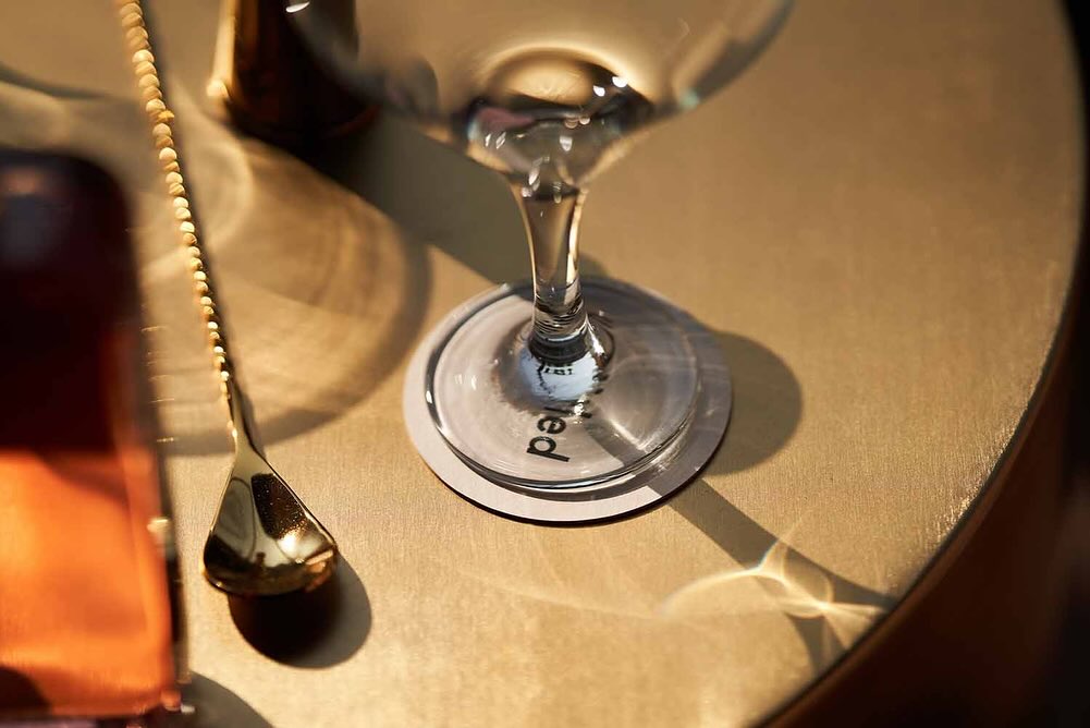 Close-up of a cocktail glass on a table with a golden spoon and a coaster reflecting light, creating an inviting ambiance.