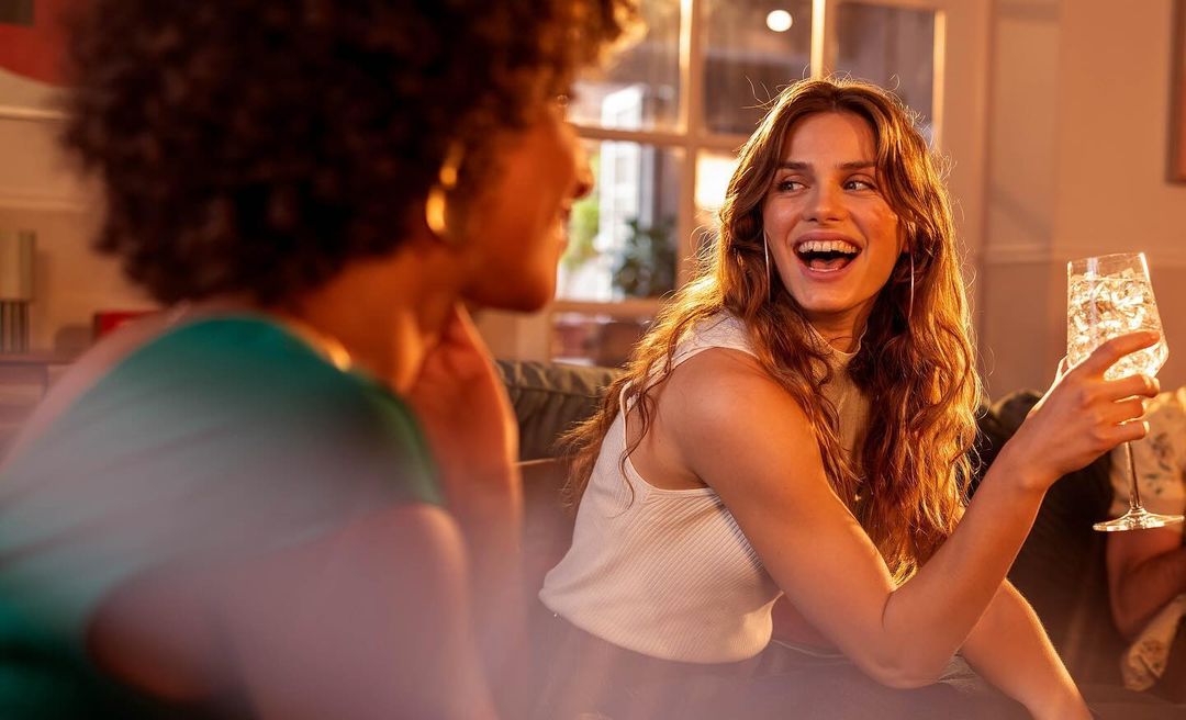 Two women socialize indoors, one holding a drink with ice. The other woman is partly visible, warm indoor lighting and casual setting.