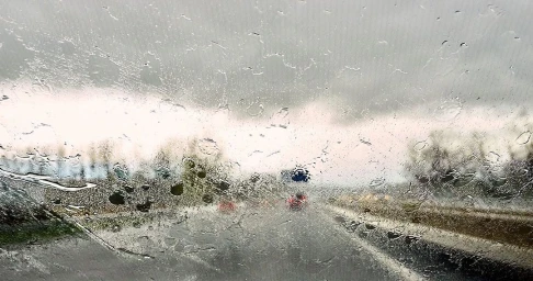 Raindrops and streaks cover a car windshield looking out onto a wet highway under an overcast sky.