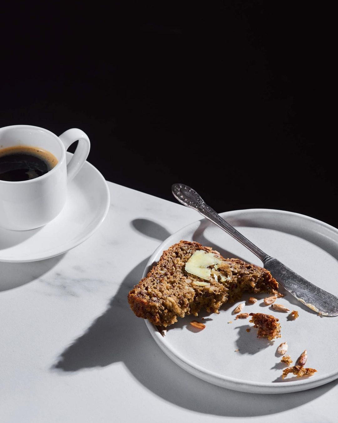 Overhead shot of a slice of banana bread with butter on a white plate with a knife, alongside a cup of black coffee on a marble surface.