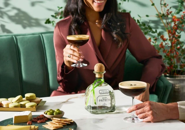 Two people enjoying espresso martinis with Patron tequila, cheese, crackers, macarons, and olives on a white marble table.