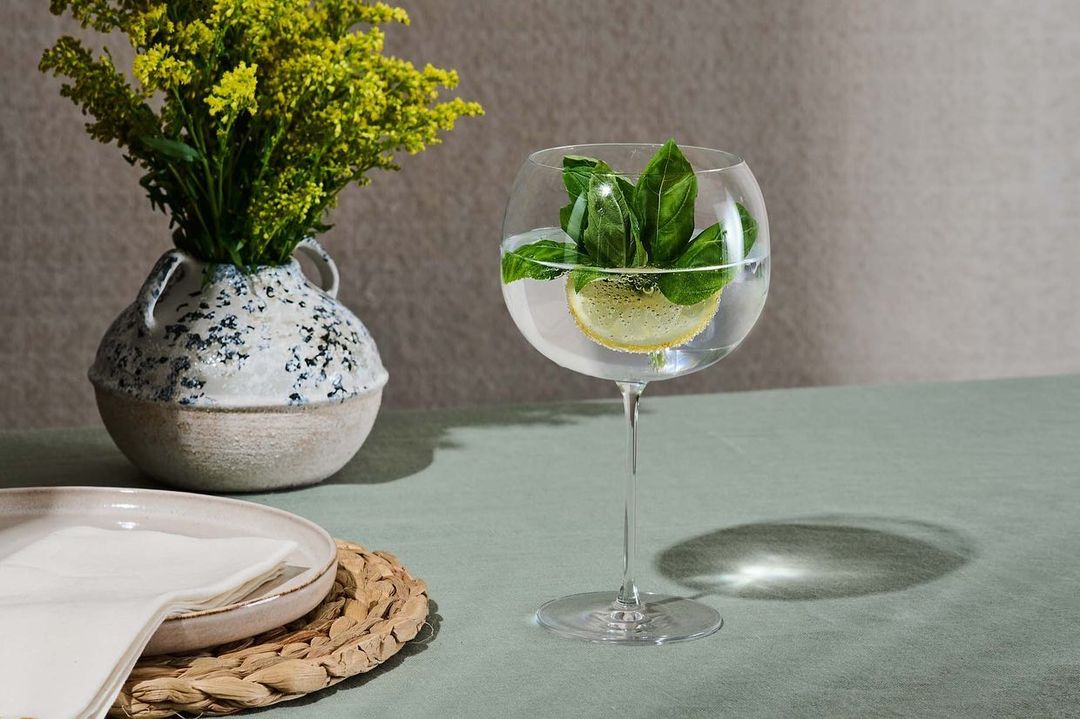 Still life food photography featuring a glass of water with lemon and basil, a flower vase, and dishes on a green table.