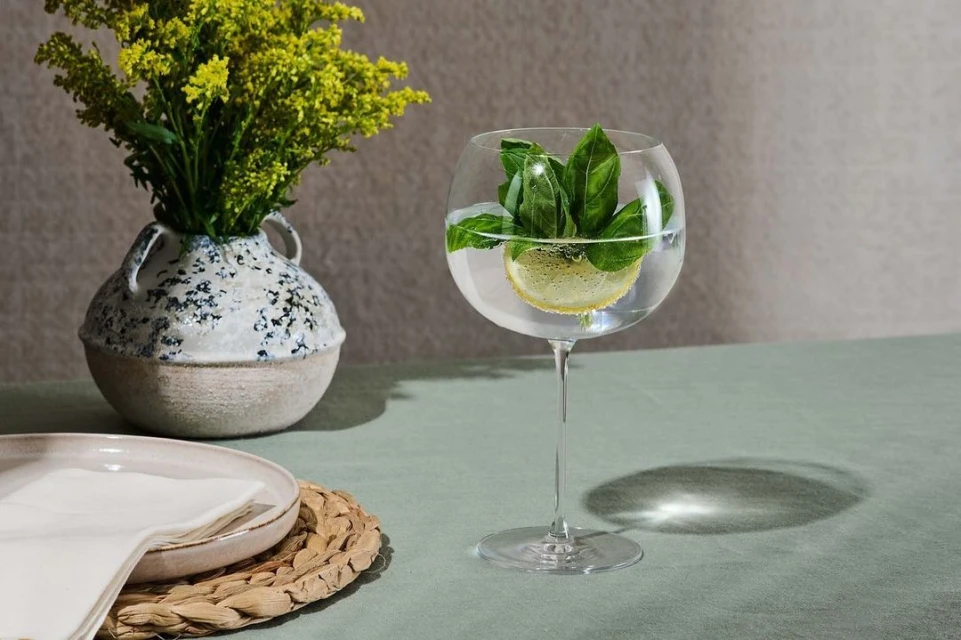 Still life food photography featuring a glass of water with lemon and basil, a flower vase, and dishes on a green table.