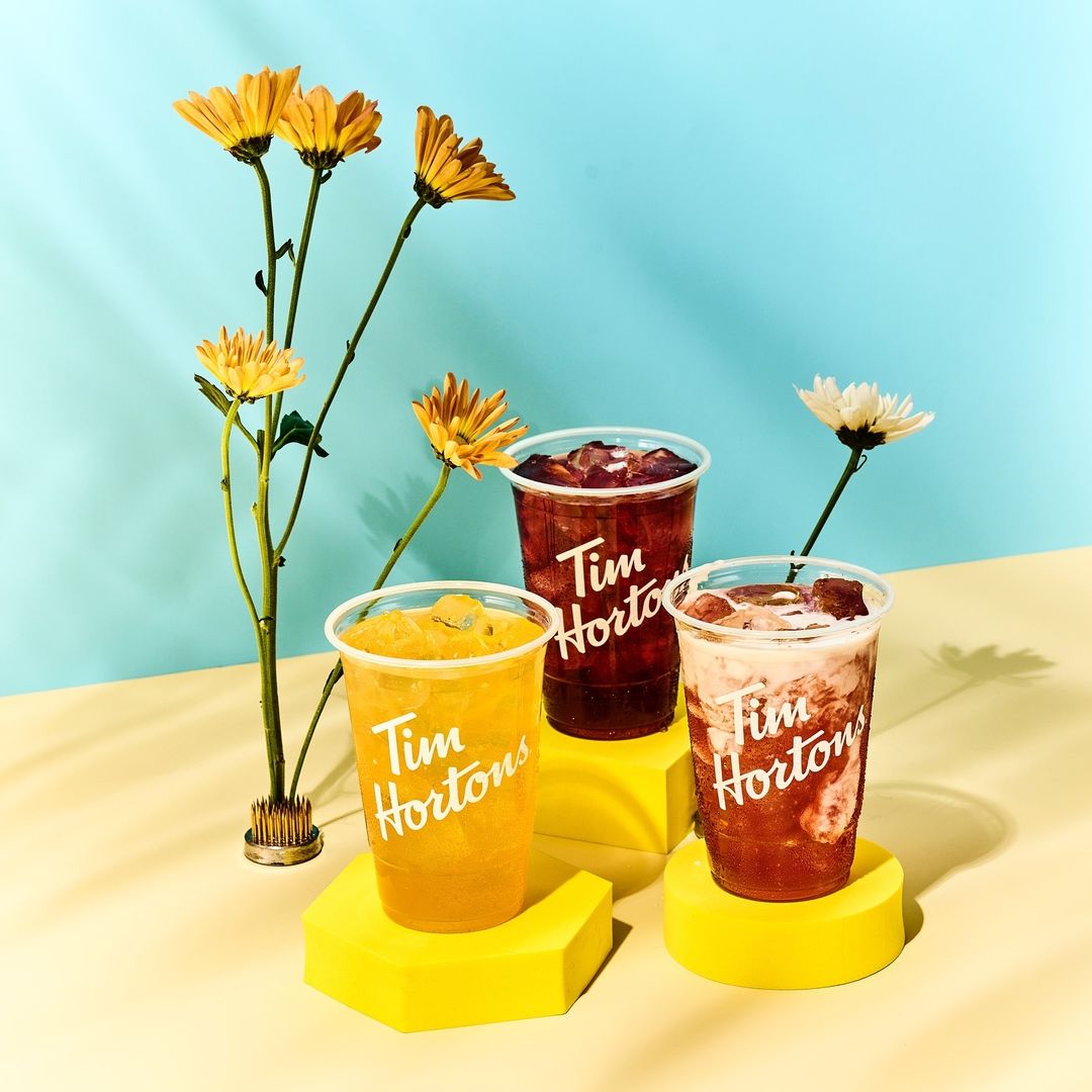 Three Tim Hortons beverages with ice displayed on yellow geometric stands with yellow and white flowers against a blue background.