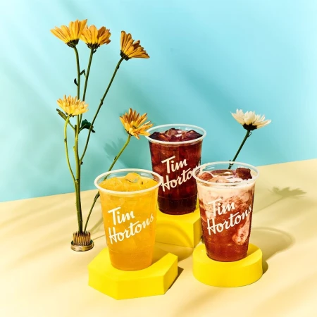 Three Tim Hortons beverages with ice displayed on yellow geometric stands with yellow and white flowers against a blue background.