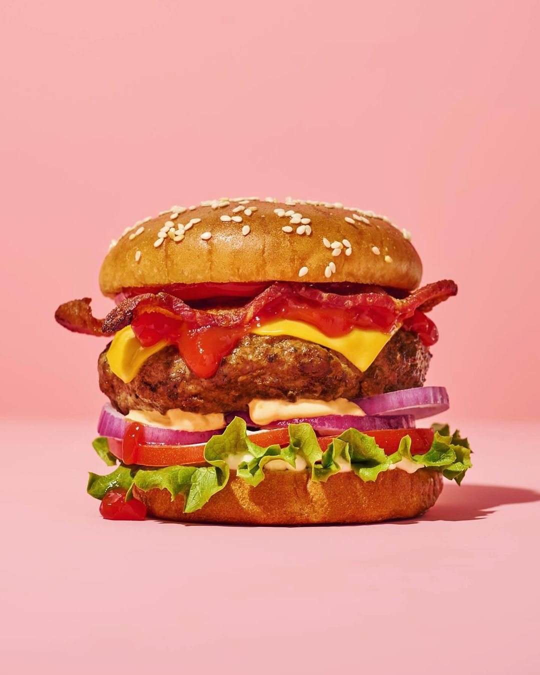 A hamburger with bacon, lettuce, tomatoes, onions and sauce on a pink background, illuminated by bright studio lighting.