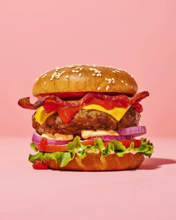 A hamburger with bacon, lettuce, tomatoes, onions and sauce on a pink background, illuminated by bright studio lighting.