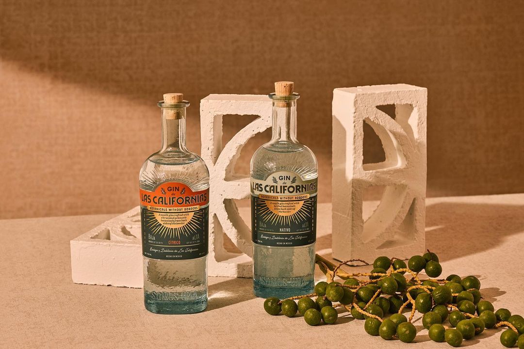 Two bottles of Las Californias gin are showcased against a neutral backdrop, accompanied by a decorative white block and a cluster of green fruits.