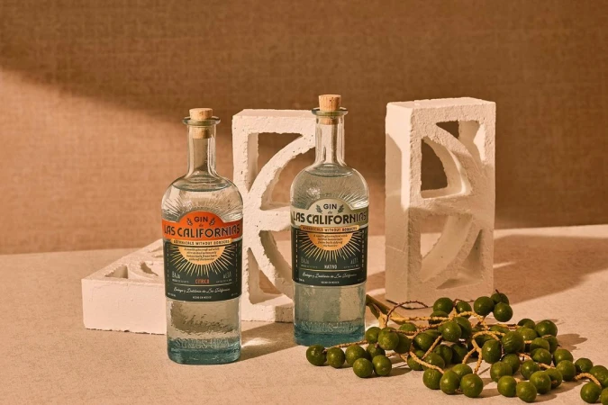 Two bottles of Las Californias gin are showcased against a neutral backdrop, accompanied by a decorative white block and a cluster of green fruits.