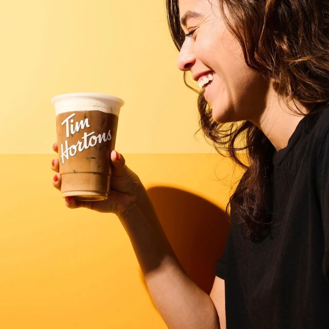 Smiling woman in black shirt holds a Tim Hortons iced coffee against yellow background. Beverage is primary focus.