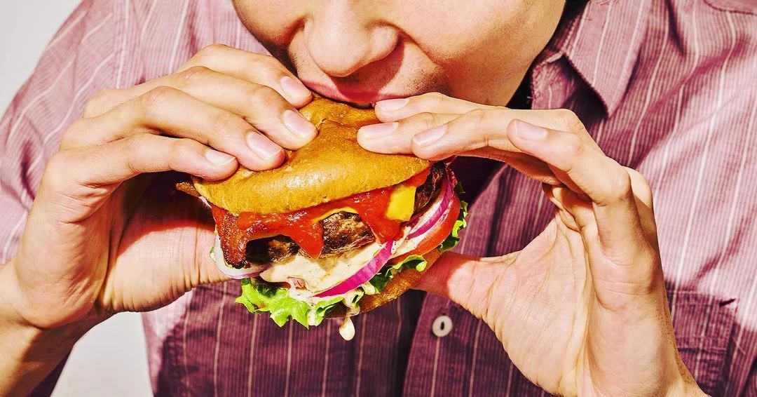 Close-up of an adult eating a large hamburger with visible toppings, showcasing textures and colors of ingredients against a white background.