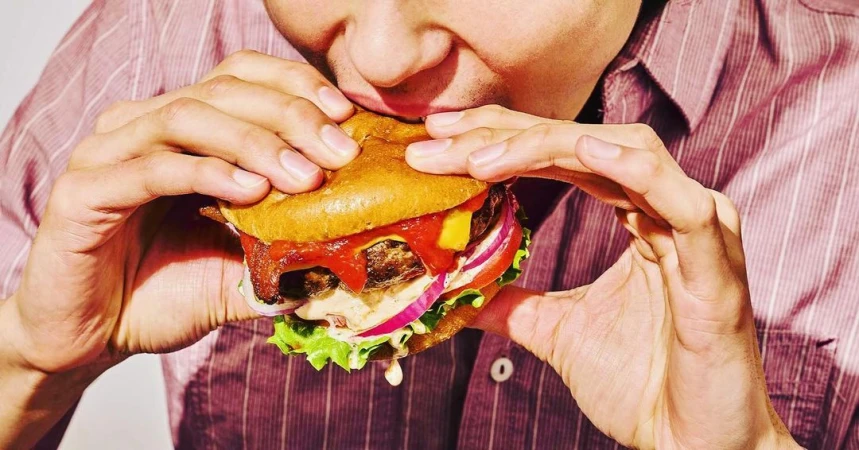 Close-up of an adult eating a large hamburger with visible toppings, showcasing textures and colors of ingredients against a white background.