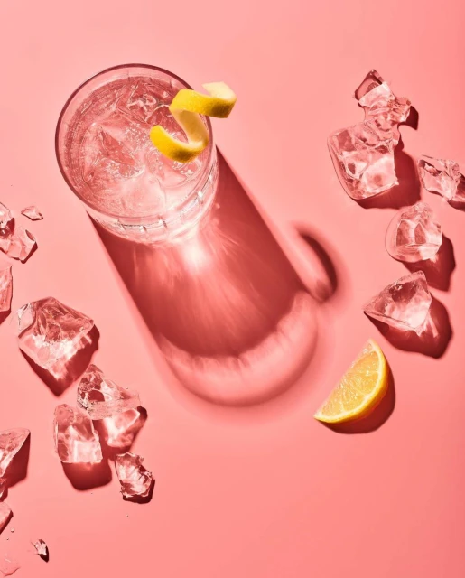 Overhead shot of a pink cocktail with lemon twist and ice chunks on a pink surface, capturing a fresh and vibrant beverage scene.