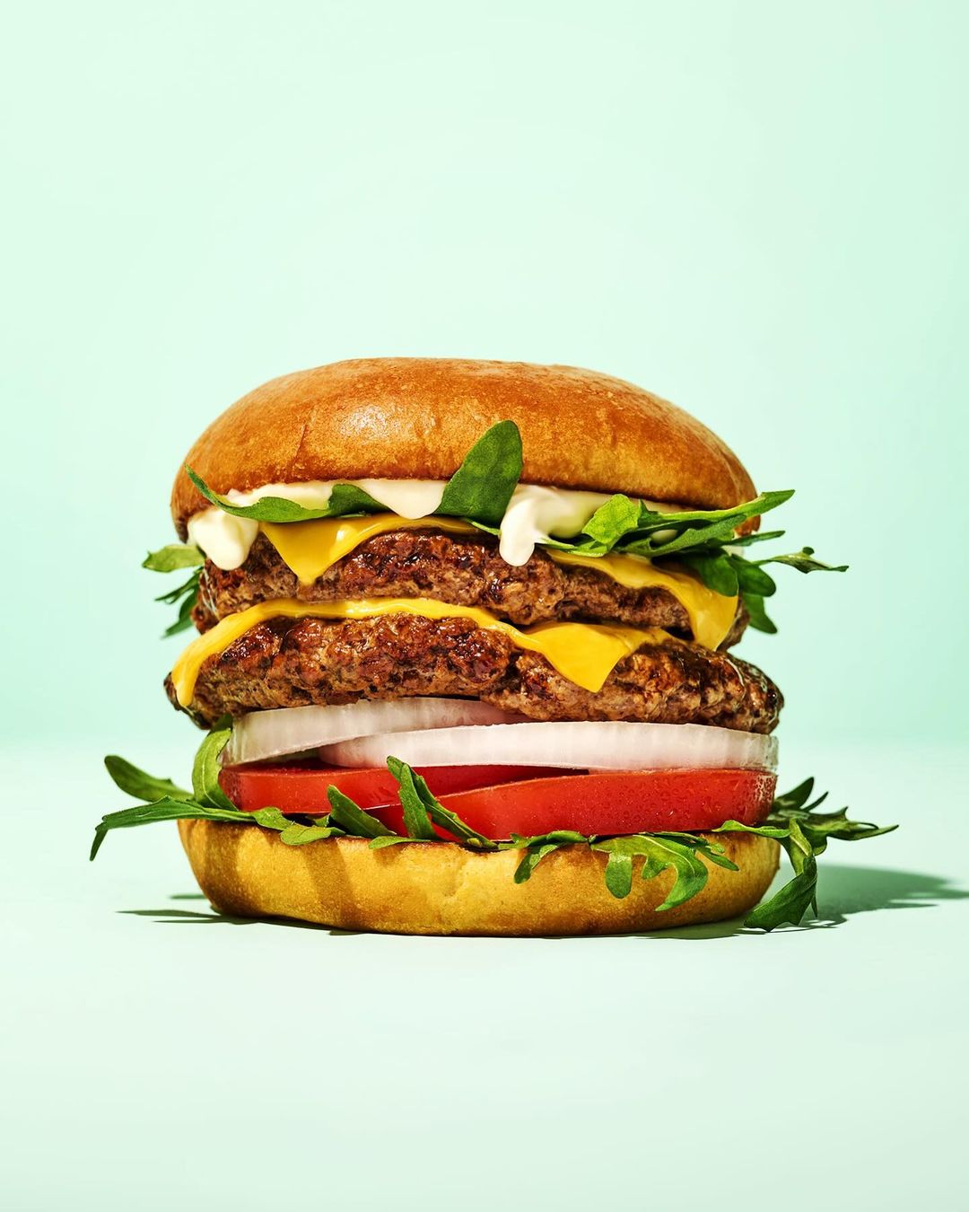 Double cheeseburger on a bun with tomato, onion, lettuce and sauce, set against a light blue background in a studio food shot.