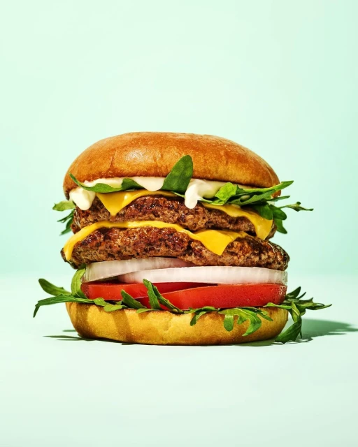 Double cheeseburger on a bun with tomato, onion, lettuce and sauce, set against a light blue background in a studio food shot.