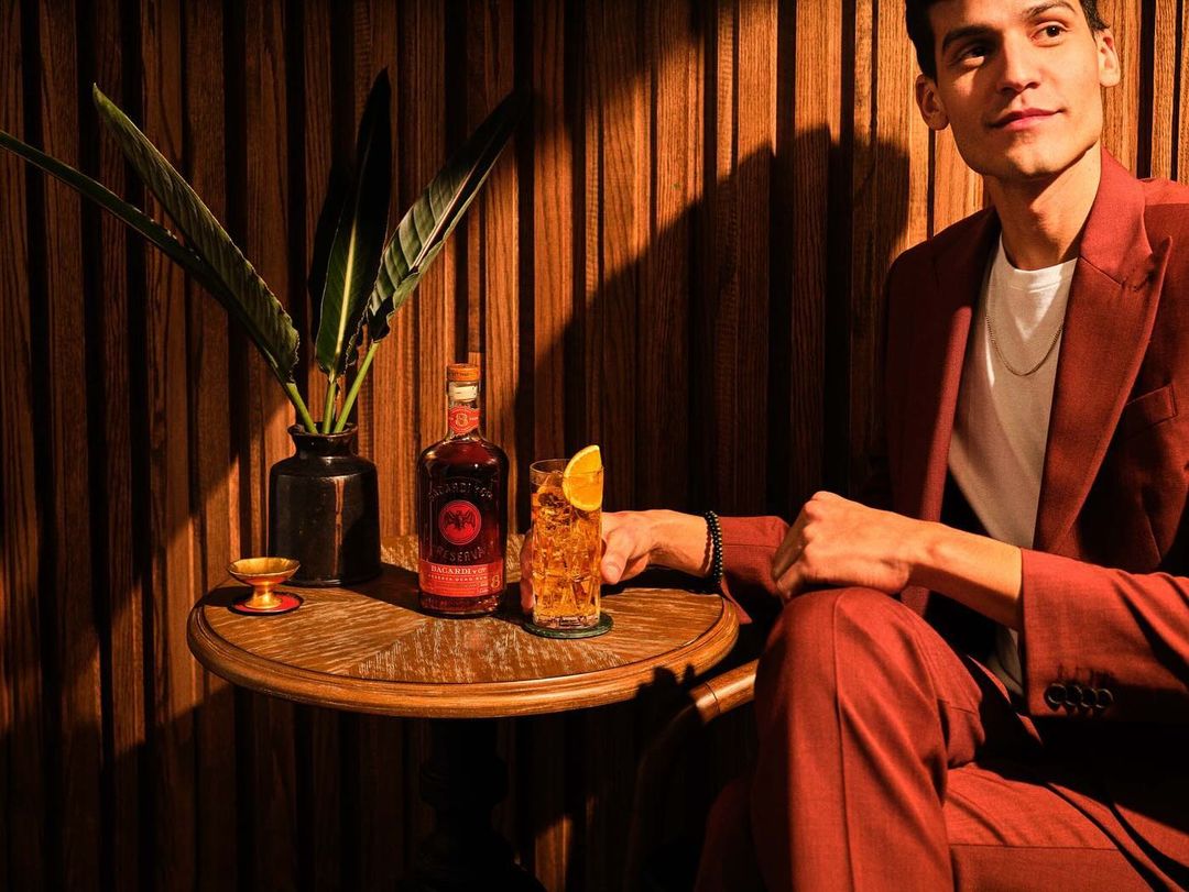 Adult male in rust-colored suit with beverage, glass, and bottle on wooden table against striped wood wall, indoor setting.