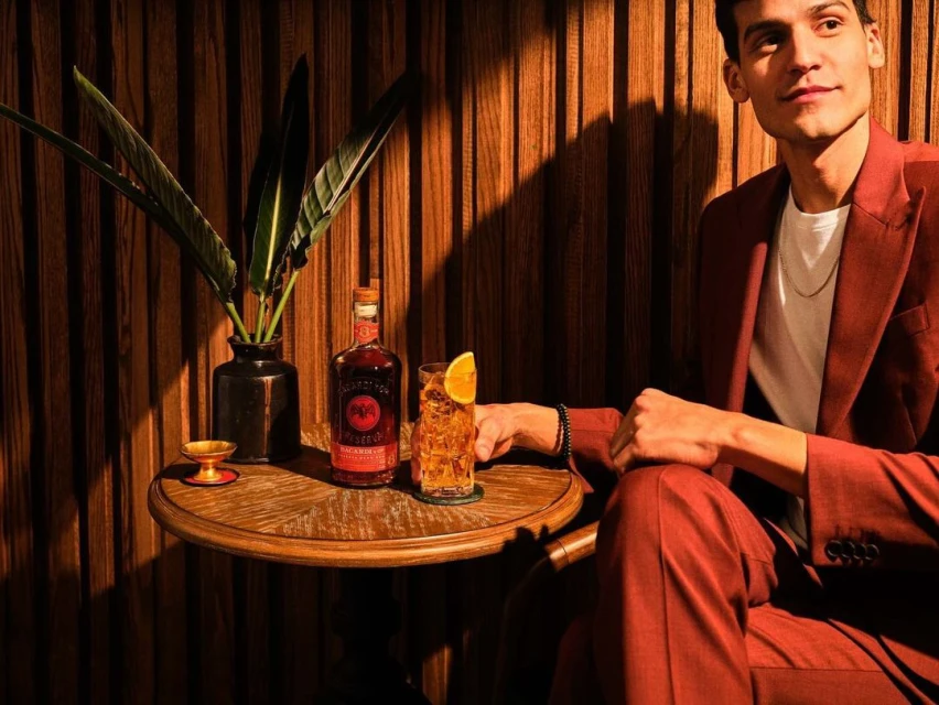 Adult male in rust-colored suit with beverage, glass, and bottle on wooden table against striped wood wall, indoor setting.