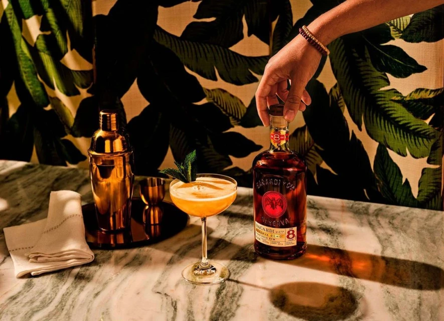 Cocktail next to Bacardi Reserva Ocho rum bottle on marble table with person's hand, gold barware, mint garnish, and tropical wallpaper.