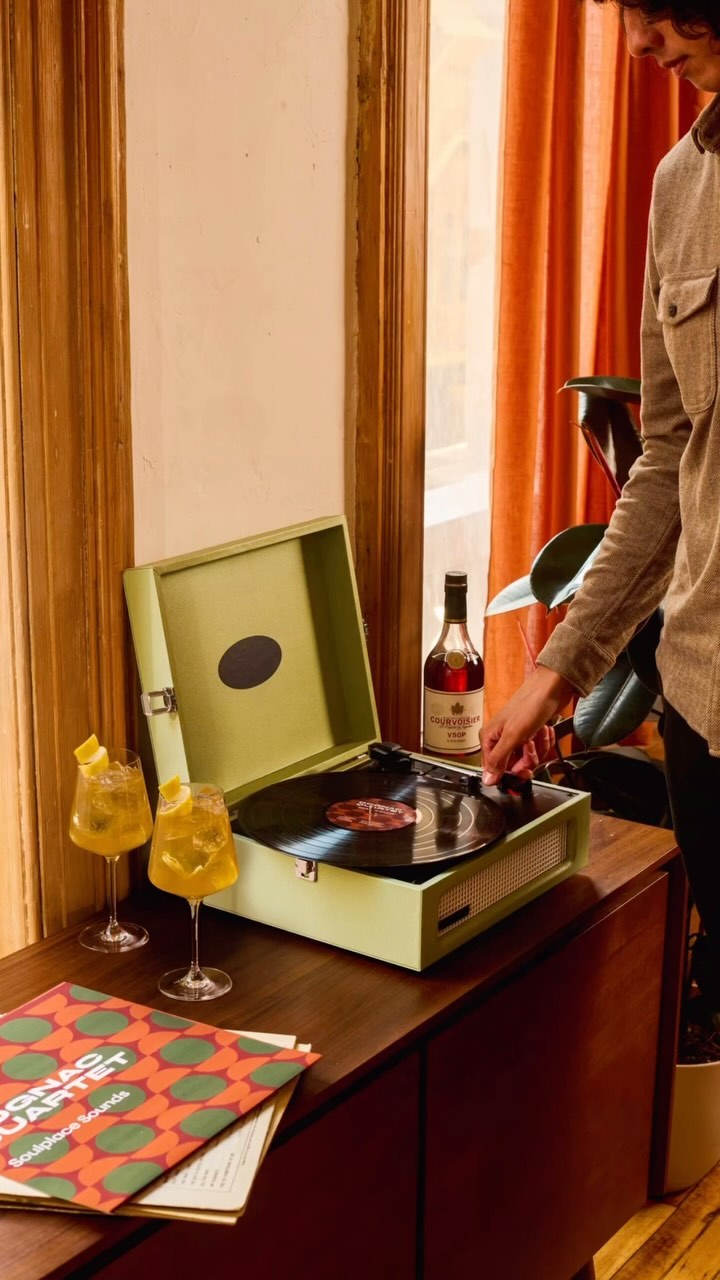 Adult reaching for vinyl record on green vintage turntable; drinks, liquor bottle, album on wood cabinet near window with orange curtains.