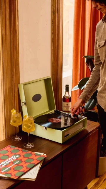 Adult reaching for vinyl record on green vintage turntable; drinks, liquor bottle, album on wood cabinet near window with orange curtains.