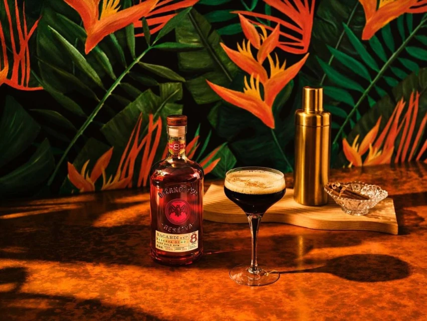 Rum bottle and cocktail with shaker and cinnamon sticks against a tropical leaf pattern background on an orange textured surface.