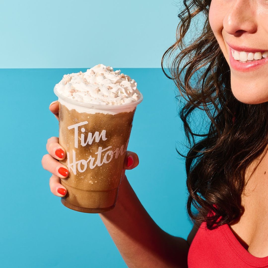Woman holds an iced cappuccino from Tim Horton's with whipped cream on top against a split blue background, showcasing a refreshing beverage.