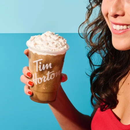 Woman holds an iced cappuccino from Tim Horton's with whipped cream on top against a split blue background, showcasing a refreshing beverage.