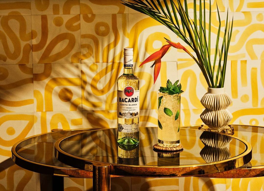 Still life of Bacardi Carta Blanca rum bottle and a mojito cocktail on a gold table against a patterned yellow background.