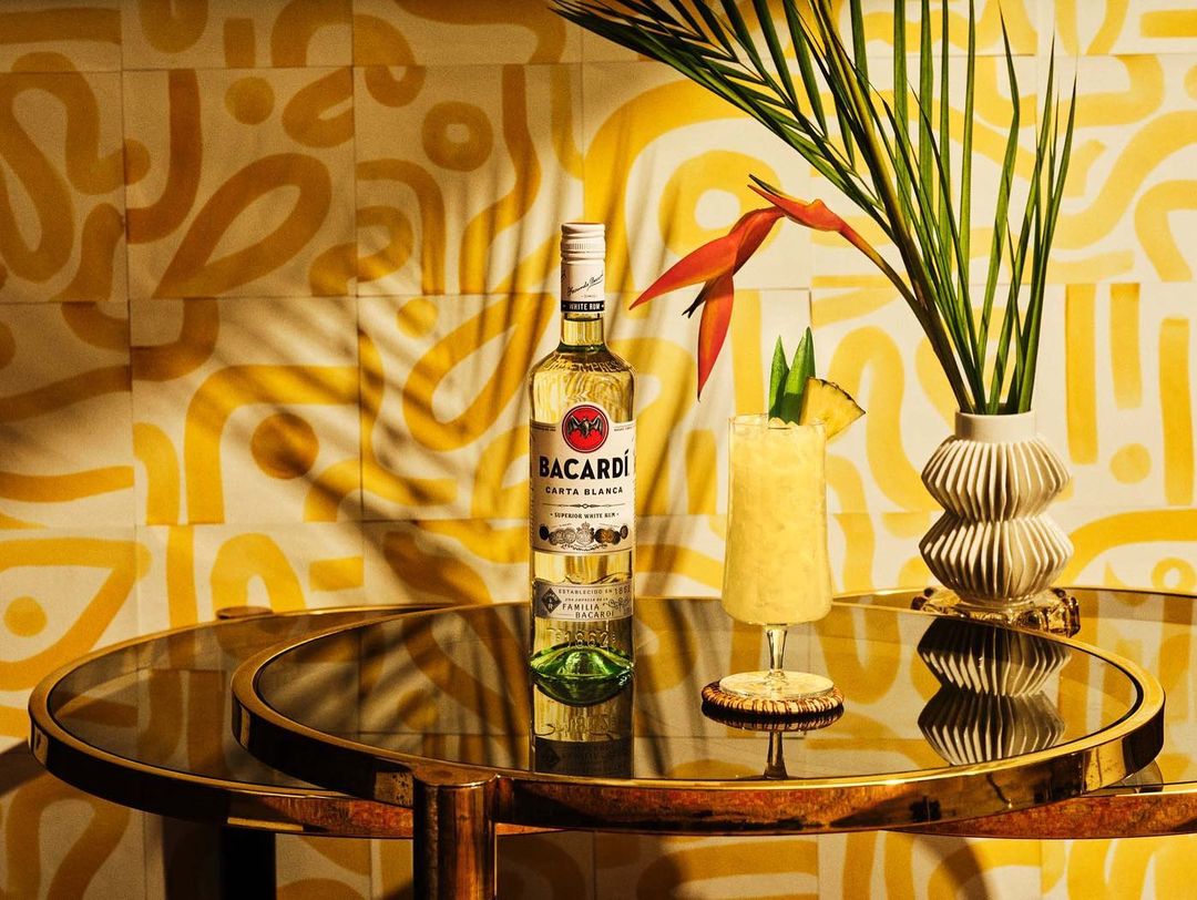 Still life shot of a bottle of Bacardi rum and a tropical cocktail on a round table, with a patterned backdrop and tropical foliage.