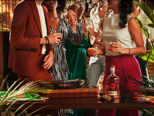 People at a party with a record player, drinks, and tropical plants, enjoying music and each other's company in a lively indoor setting.