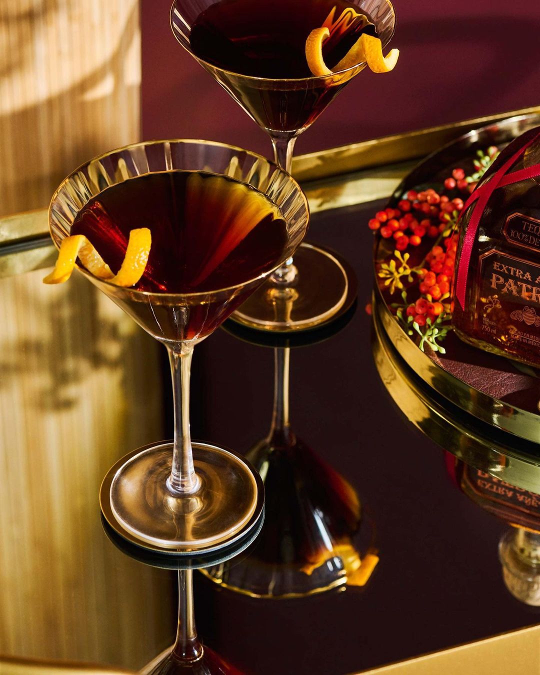 Two cocktails with orange twists on a reflective gold tray in a dimly lit setting, next to a bottle with red berries.