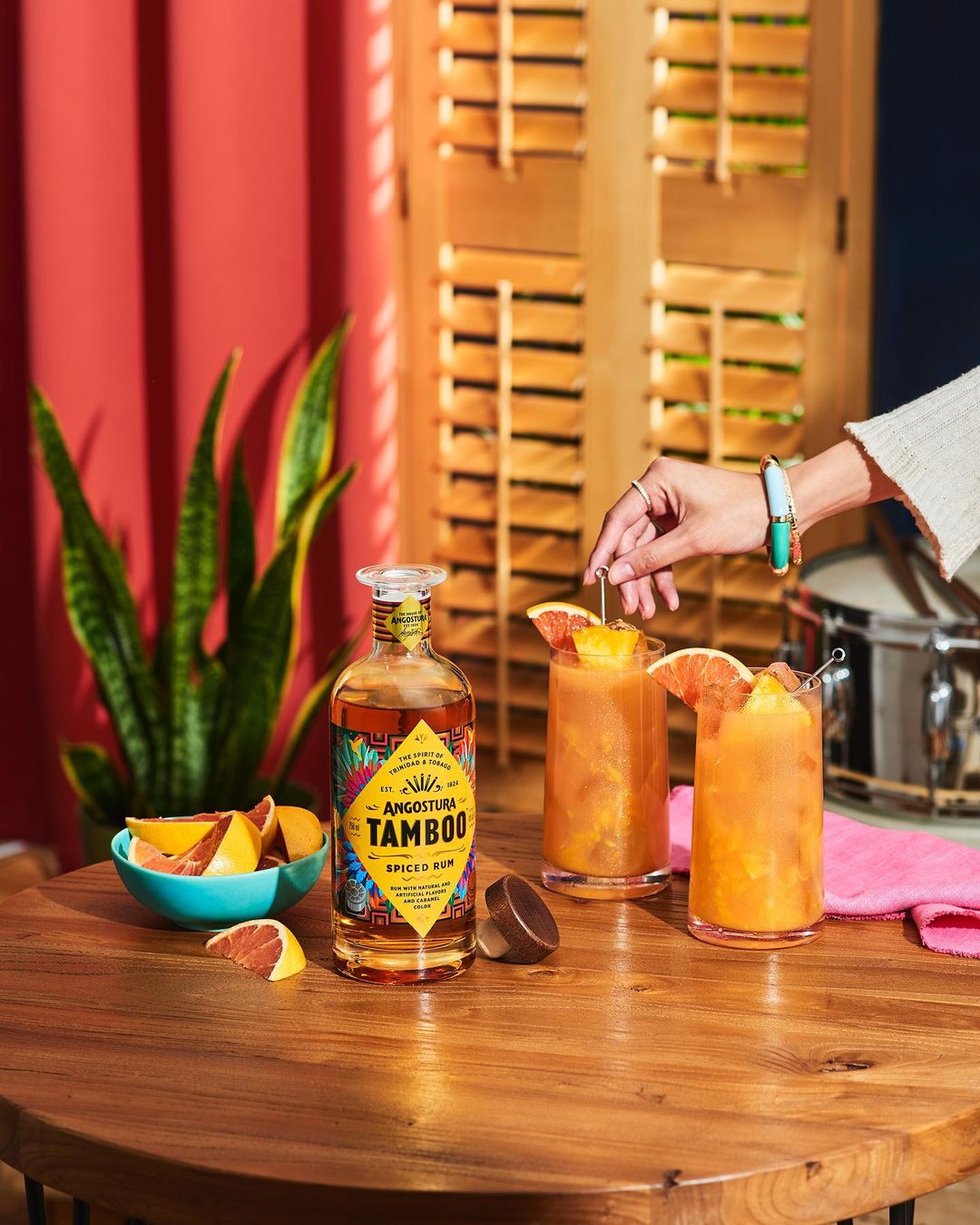 Angostura Tamboo spiced rum bottle on wooden table with cocktails, grapefruit slices, and decorative shutters in background.