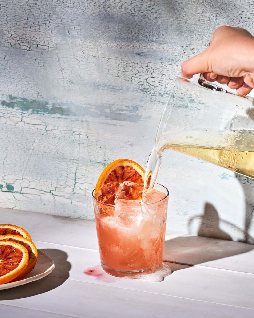 A person pours a cocktail from a pitcher into a glass filled with ice and an orange slice, with additional orange slices nearby.