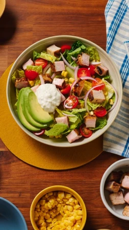 Overhead shot of a vibrant salad with grilled pork, avocado, and corn, arranged in a white bowl on a yellow and wood background.